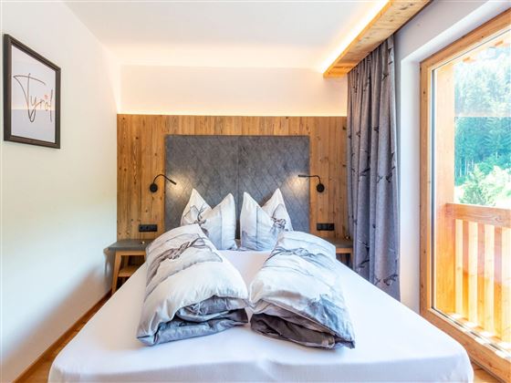 A bright and modern bedroom with a large bed and stylish pillows. The window offers a view of nature outside.