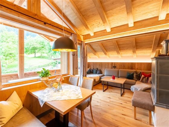 A cozy living space with a wooden ceiling and large windows. The room is bright and inviting, with a table and comfortable seating.