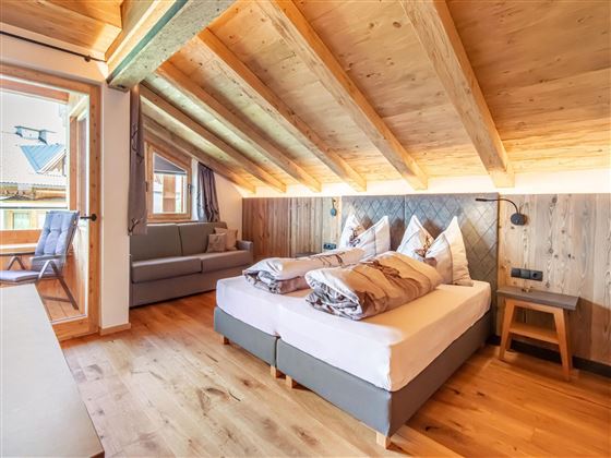 A cozy bedroom with wooden walls and a sloped ceiling. There is a double bed, a sofa, and large windows that let in a lot of light.