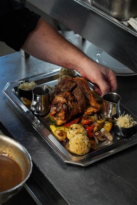 Un piatto servito con un grande pezzo di carne, accompagnato da verdure varie e contorni. Una mano si allunga verso il piatto, che si trova su un tavolo in una cucina di ristorante.