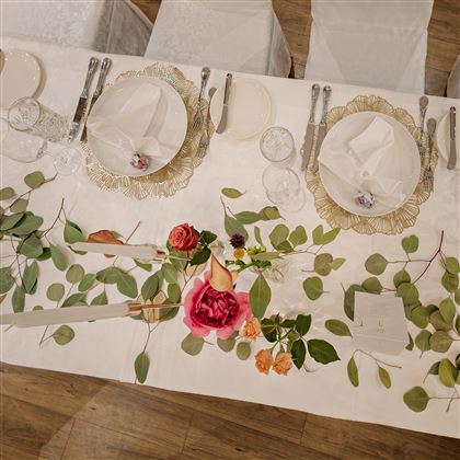 Una tavola elegantemente apparecchiata con piatti bianchi e posate argentate. La decorazione del tavolo include fiori freschi e foglie verdi.