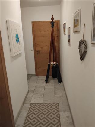 Een lichte gang met een houten deur en verschillende foto's aan de muren. In het midden staat een kledingrek met een zwart en een grijs kledingstuk.