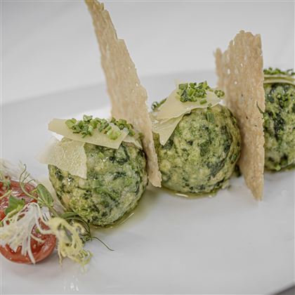 Tre gnocchi verdi con formaggio e un chip croccante su un piatto bianco. Accanto ci sono pomodorini e erbe fresche.