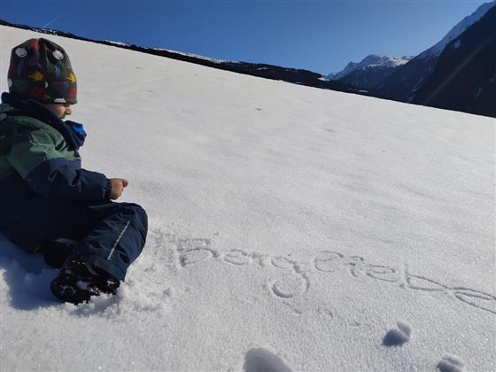 Een kind zit in de sneeuw en kijkt naar de bergen. Op de sneeuw staat de tekst "BergLiefde".