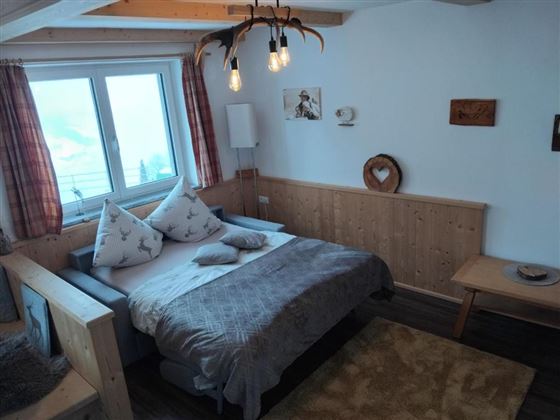 Een gezellige slaapkamer met een groot bed, zachte kussens en houten muren. Grote ramen laten veel daglicht binnen en geven uitzicht naar buiten.