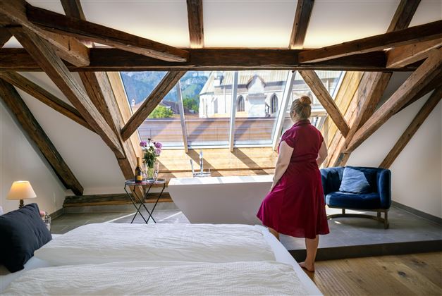 A cozy bedroom with wooden beams and large windows. A woman stands in front of the view and looks out at a beautiful landscape.