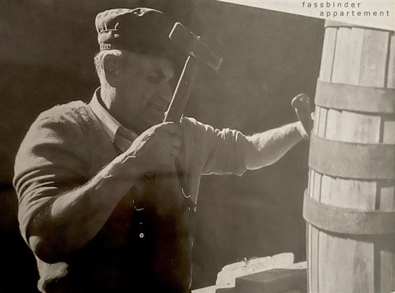 A man is working with a hammer on a barrel. He is wearing a cap and has a concentrated expression.