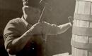 A man is working with a hammer on a barrel. He is wearing a cap and has a concentrated expression.