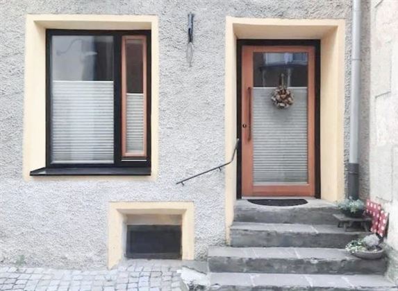 A front door in a gray building with windows and a small staircase. Plants in pots are on the ground and a wreath is hanging on the door.