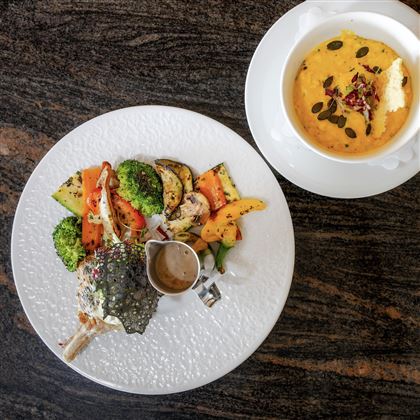 Un bel piatto con verdure grigliate e filetto di pesce, servito con una piccola salsa. Accanto c'è una ciotola con una crema di zuppa.