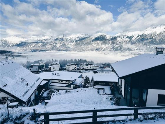 Un paysage enneigé avec des maisons au premier plan. À l'arrière-plan, on peut voir des montagnes majestueuses sous un ciel bleu nuageux.
