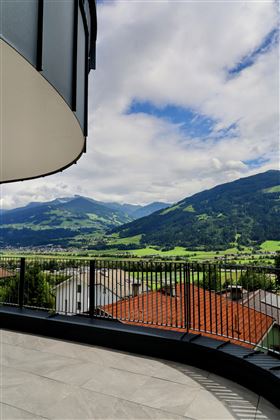Een mooi uitzicht vanaf een terras op groene bergen en wolken aan de hemel. Het landschap is rustig en uitnodigend, met zachte heuvels en charmante huizen op de voorgrond.