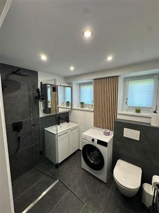 Un moderno bagno con una doccia, lavatrice e wc. Le pareti sono decorate con piastrelle scure e ci sono luci a soffitto chiare.