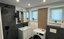 Un moderno bagno con una doccia, lavatrice e wc. Le pareti sono decorate con piastrelle scure e ci sono luci a soffitto chiare.