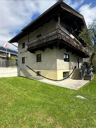 Una tradizionale casa a due piani con un balcone in legno. Il prato è verde e curato, e si possono vedere alcuni alberi e piante.