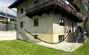 Una tradizionale casa a due piani con un balcone in legno. Il prato è verde e curato, e si possono vedere alcuni alberi e piante.