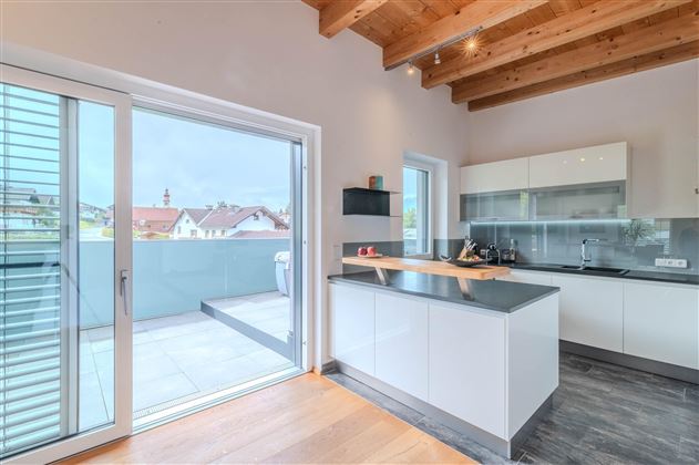 A modern kitchen with bright walls and wooden ceilings. The room has a large window front and access to a terrace.