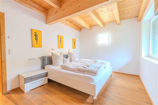 A modern bedroom with a double bed and wooden ceilings. Yellow pictures hang on the walls, and there is a window that lets in a lot of light.