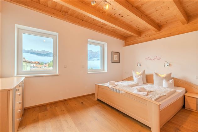 A bright bedroom with a wooden ceiling and large windows. The view of the mountains and the lake creates a relaxing atmosphere.