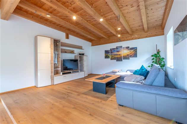 A modern living room with a wooden beam ceiling. The furnishings include a gray couch, a coffee table, and a TV unit.