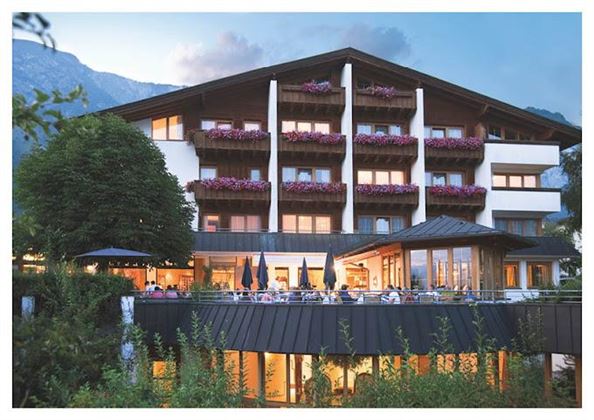 A charming hotel with balconies adorned with flowers. It is situated in a beautiful setting, surrounded by trees and mountains.
