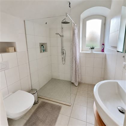 Ein modernes Badezimmer mit einer Duschkabine, einer Toilette und einem Waschbecken. Die Wände sind hell gefliest und ein Fenster sorgt für Tageslicht.