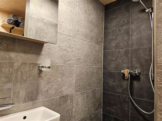 Ein modernes Badezimmer mit grauen Fliesen und einer Dusche. Es gibt ein Waschbecken und einen Spiegel mit Handtüchern im Regal.