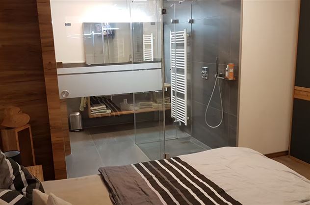 A modern bedroom with an open bathroom area. The shower is separated by glass walls, and the room has a simple, elegant decor.