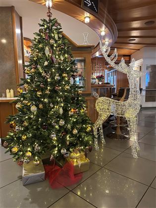 A festively decorated Christmas tree with shiny ornaments and lights stands next to a glowing reindeer. The tree is adorned with gifts and creates a cozy atmosphere.