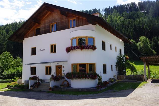 A charming two-story house surrounded by trees and flowers. The facade is white with wood cladding and blooming balcony plants.