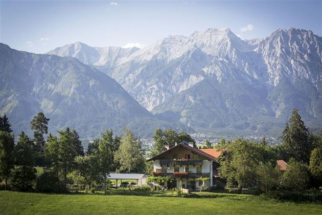 A picturesque house in a green landscape with majestic mountains in the background. The sky is clear and nature is lush and peaceful.