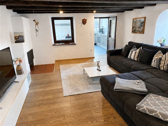 Ein gemütliches Wohnzimmer mit einem großen, grauen Sofa und einem flachen Fernseher. Der Raum hat Holzbalken an der Decke und einen warmen Holzboden.