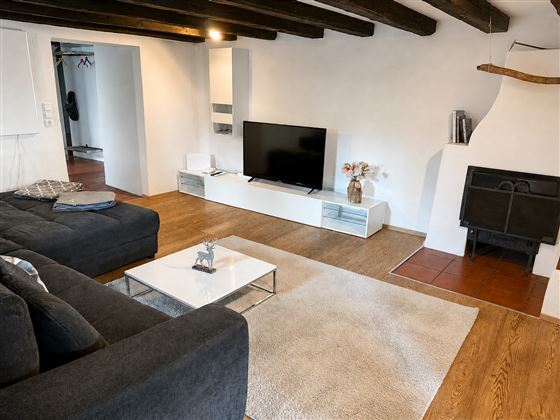 Ein modernes Wohnzimmer mit einer großen Couch und einem Fernseher. Der Raum hat Holzdeckenbalken und einen Kamin, der für Gemütlichkeit sorgt.