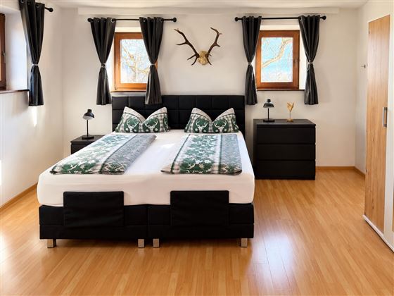 Ein modernes Schlafzimmer mit einem Doppelbett und grüner Bettwäsche. Die Wände sind hell, und es gibt Holzfenster mit Vorhängen.