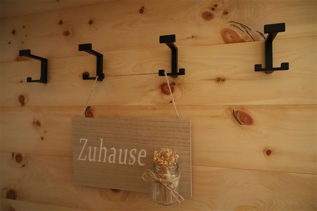Ein Holzbrett mit der Aufschrift „Zuhause“ hängt an einer Wand mit schwarzen Haken. Daneben befinden sich drei schwarze Haken, die für Jacken oder Dekorationen genutzt werden können.