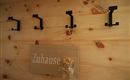 Ein Holzbrett mit der Aufschrift „Zuhause“ hängt an einer Wand mit schwarzen Haken. Daneben befinden sich drei schwarze Haken, die für Jacken oder Dekorationen genutzt werden können.