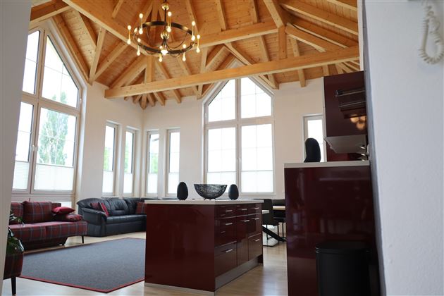 Ein modernes Wohnzimmer mit hohen Fenstern und Holzdecken. Es gibt eine offene Küche mit roten Möbeln und komfortablen Sitzmöglichkeiten.