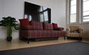 Ein gemütliches Wohnzimmer mit einem rotkarierten Sofa und einem kleinen Korbstuhl. Im Hintergrund befindet sich ein großer Fernseher und eine Pflanze steht neben dem Sofa.