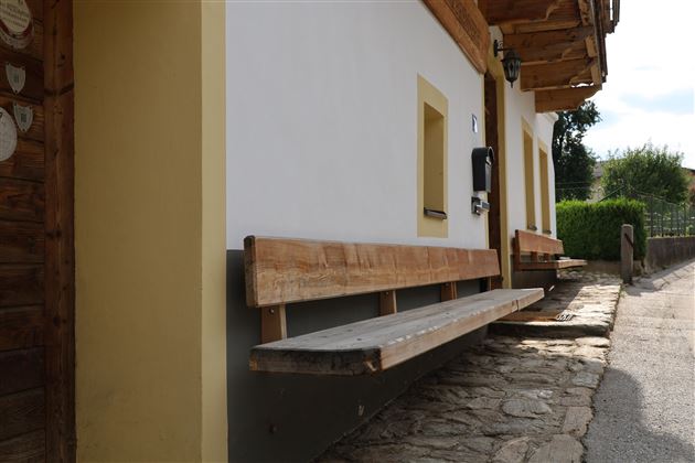 Eine Holzbank vor einem Haus mit einer weißen Wand. Im Hintergrund sind Pflanzen und ein gepflasterter Weg zu sehen.