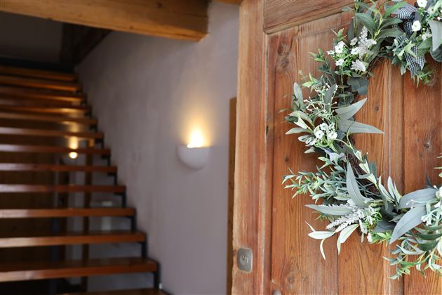 Eine einladende Tür mit einem schönen Kranz aus Pflanzen und einem Holzgeländer im Hintergrund. Die Treppe führt nach oben und ist in warmem Licht beleuchtet.