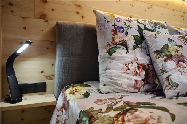 Ein gemütliches Schlafzimmer mit einem Bett, das mit floralem Bettzeug und Kissen bedeckt ist. Neben dem Bett steht eine moderne Leselampe auf einem Nachttisch.