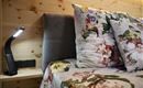 Ein gemütliches Schlafzimmer mit einem Bett, das mit floralem Bettzeug und Kissen bedeckt ist. Neben dem Bett steht eine moderne Leselampe auf einem Nachttisch.