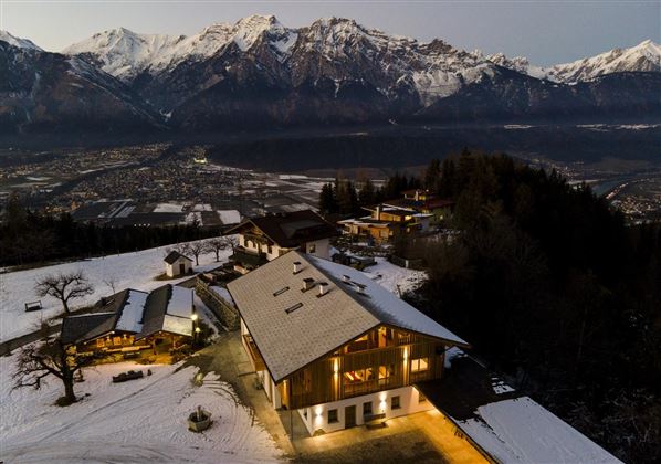 A picturesque mountain landscape with snow-covered peaks in the background. Below, there is a modern wooden house that is softly illuminated.