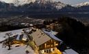 A picturesque mountain landscape with snow-covered peaks in the background. Below, there is a modern wooden house that is softly illuminated.