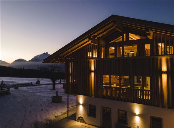 A modern wooden house at dusk, illuminated by warm light. Snow-covered mountains are visible in the background.
