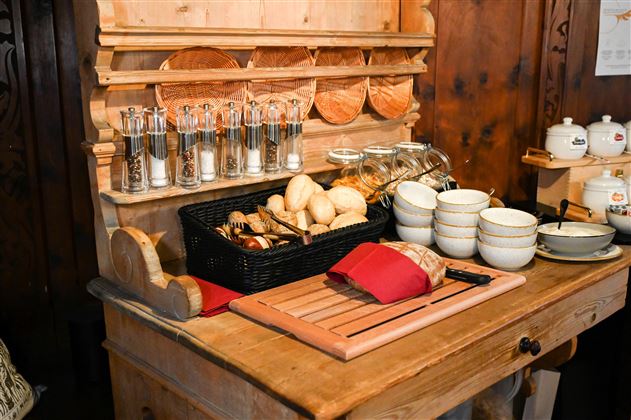 A rustic wooden counter with spice containers, a bread basket, and dishes. On the work surface lies a cutting board and a red cloth.
