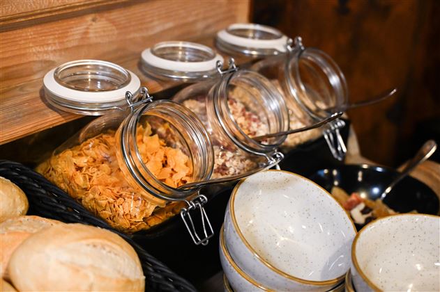 A selection of cereals in jars, standing on a wooden shelf. Next to them are bowls and rolls.