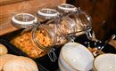 A selection of cereals in jars, standing on a wooden shelf. Next to them are bowls and rolls.
