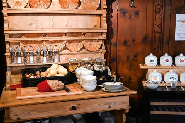 A rustic wooden cupboard with various glasses, bowls, and plates. On the workspace, fresh rolls and ingredients for a meal are arranged.