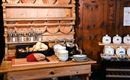 A rustic wooden cupboard with various glasses, bowls, and plates. On the workspace, fresh rolls and ingredients for a meal are arranged.